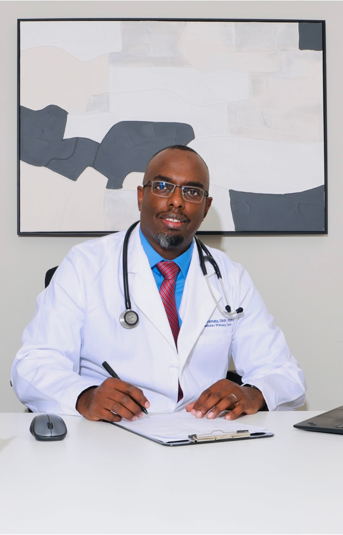 Dr. Joseph L. Giramata, DNP, FNP-C, seated at a desk in a white coat with a stethoscope